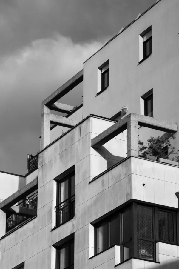 Daniel · 1961 x 2942 Cette photographie en noir et blanc met en scène un immeuble moderne aux lignes géométriques marquées. Le sujet principal est l’architecture, avec ses balcons asymétriques et ses jeux de volumes. La composition joue sur les contrastes entre pleins et vides, accentués par des angles saillants.Le cadrage serré isole une partie de la façade, créant une ambiance urbaine et minimaliste. La lumière, naturelle et douce, souligne les textures et les ombres, renforçant le côté épuré. Les tons dominants, en niveaux de gris, accentuent la sobriété et la modernité du style architectural. L’ambiance est à la fois froide et élégante, reflétant une esthétique contemporaine.