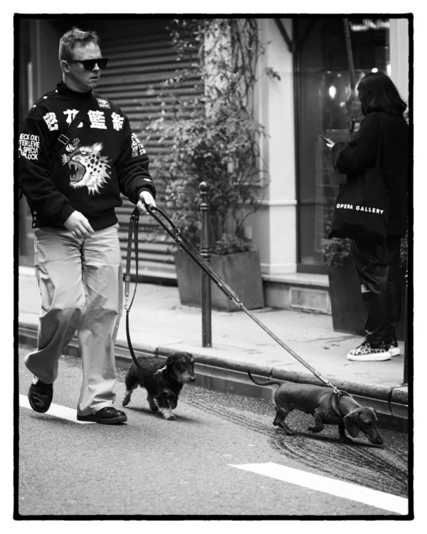 Thierry · 3072 x 3840 Cette photographie en noir et blanc met en scène un homme, sujet principal, vêtu d'une veste militaire, promenant deux chiens avec une laisse. La composition est simple et centrée, avec un cadrage serré sur le sujet et son environnement immédiat. L’ambiance est urbaine, avec des bâtiments et des passants en arrière-plan.La lumière semble naturelle, probablement en journée, créant des contrastes marqués. Le style est documentaire et réaliste, capturant un moment du quotidien. Les tons dominants sont les gris et noirs, renforçant l’aspect authentique et intemporel de la scène.