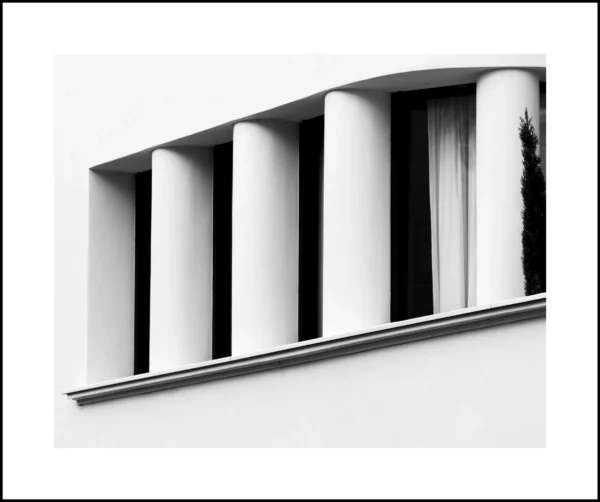 Thierry · 21/02/2026 · 4680 x 3912 Cette photographie met en scène une façade architecturale minimaliste avec des colonnes verticales alignées, encadrées par un rebord horizontal. Le sujet principal est cette structure géométrique épurée.La composition repose sur des lignes droites et une symétrie presque parfaite. Le cadrage est serré, centré sur les colonnes, créant une impression de profondeur et de perspective.En noir et blanc, la lumière est douce et uniforme, accentuant les contrastes entre ombres et lumières. L’ambiance est sobre, presque austère, évoquant un style architectural classique revisité dans un esprit moderne et épuré.
