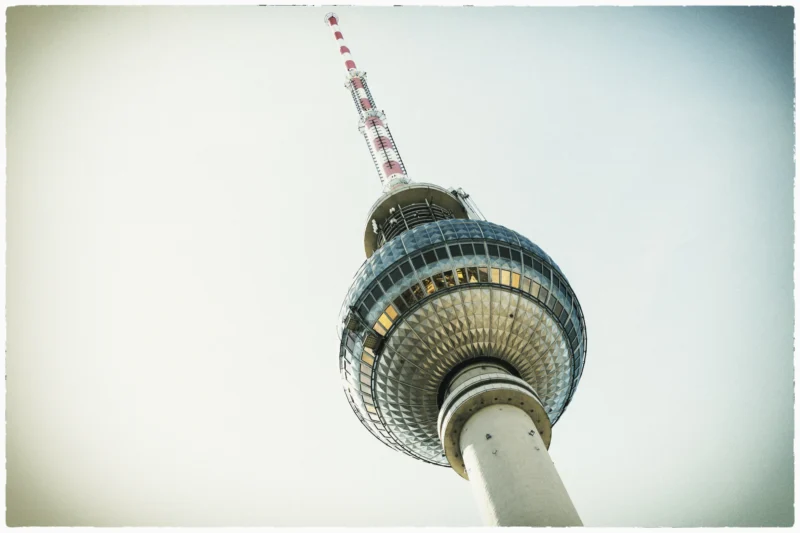 Cette photographie met en scène la **Tour de télévision de Berlin**, sujet principal imposant. Le cadrage est serré, centré sur la structure métallique et son sommet arrondi, avec une légère inclinaison vers le haut.La lumière est douce, légèrement voilée, créant une ambiance atmosphérique et presque nostalgique. Les couleurs dominantes sont des teintes **bleutées et grises**, adoucies par un fond flou et pâle.Le style est épuré, presque minimaliste, avec un rendu légèrement vintage. L’ambiance est **moderne et industrielle**, renforcée par les lignes architecturales géométriques.