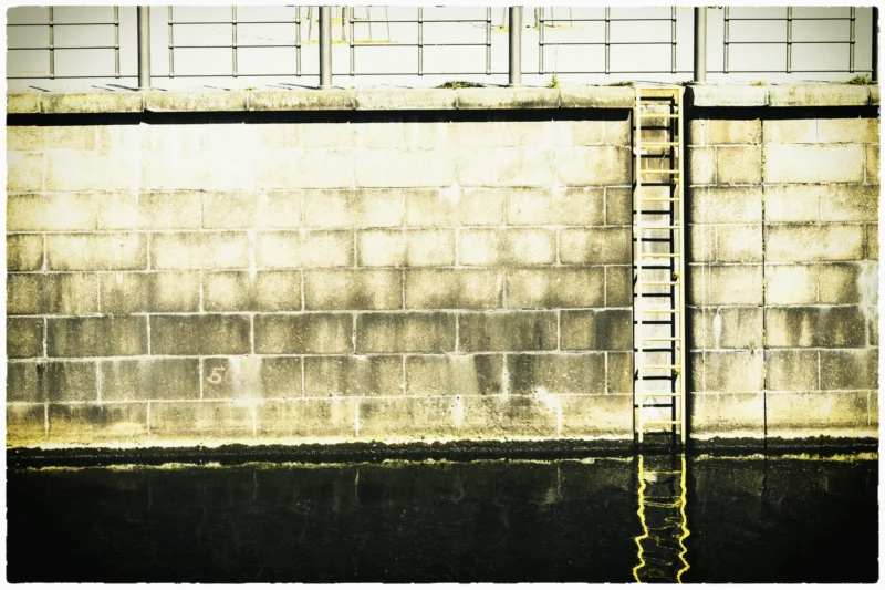 Cette photographie montre un mur en briques apparentes et une structure métallique verticale rouillée. Le sujet principal est ce mur vieillissant, marqué par le temps et l’usure.La composition est épurée, avec un cadrage serré sur la texture des briques et des traces de corrosion. La lumière semble naturelle mais tamisée, créant des contrastes doux. Les couleurs dominantes sont les tons gris-beige des briques et les reflets oxydés du métal.Le style est documentaire et brut, évoquant une ambiance industrielle désuète et mélancolique, presque nostalgique.