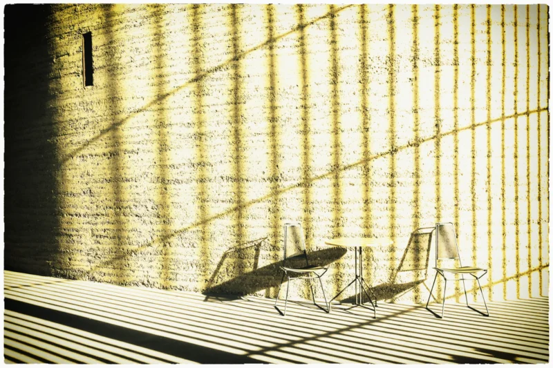 Cette photographie met en scène des **chaises transparentes** disposées sur une surface en bois, projetant des ombres allongées. La **composition** est épurée, avec un cadrage serré sur les objets et leur reflet. La **lumière**, filtrée par des **lattes verticales**, crée des jeux d'ombres et de clarté, accentuant la texture du bois et des chaises.Les **couleurs dominantes** sont des tons **jaunes sépia et bruns**, donnant un aspect vintage. Le style, **minimaliste et graphique**, évoque une ambiance **sereine et intemporelle**, presque onirique, renforcée par les contrastes lumineux.