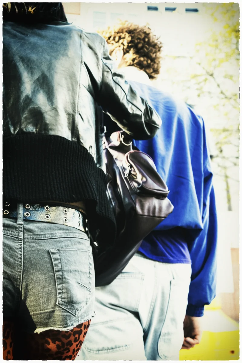 Cette photographie montre deux personnes vues de dos, formant le sujet principal. La composition met en avant leurs silhouettes, avec un cadrage serré sur leurs épaules et leurs sacs.La lumière semble naturelle, légèrement tamisée, créant des contrastes doux. Les couleurs dominantes sont des tons neutres : bleu, gris et noir, avec des touches de blanc et de rouge (chaussettes).Le style est sobre et documentaire, évoquant un moment du quotidien. L’ambiance est calme et un peu mélancolique, renforcée par les teintes sépia et le flou des contours.