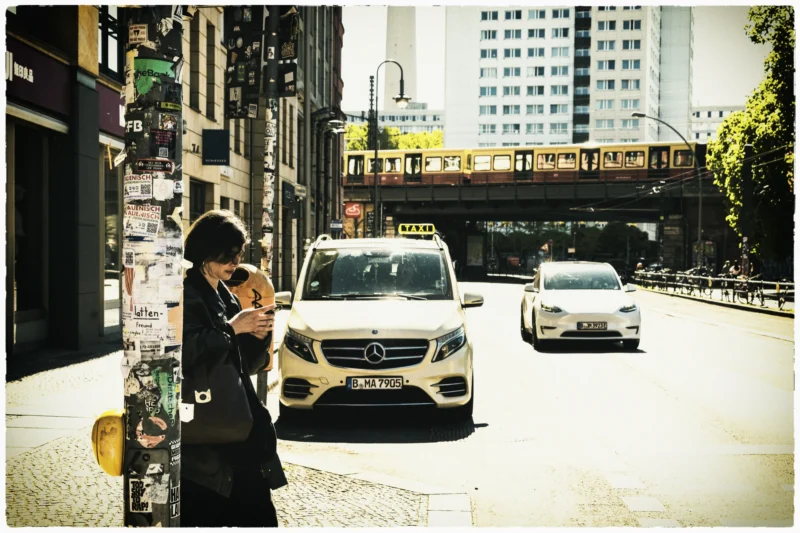 Cette photographie met en scène une femme en silhouette, de dos, tenant un téléphone, comme absorbée par son écran. La composition joue sur les contrastes entre elle et l’animation urbaine derrière elle : voitures, bâtiments et un pont surplombant.Le cadrage est serré sur la femme, avec une profondeur de champ qui floute légèrement l’arrière-plan. La lumière est naturelle, douce et légèrement dorée, évoquant un moment en fin de journée. Les couleurs dominantes sont des tons neutres (gris, beige) avec des éclats de blanc et de noir, renforçant une ambiance moderne et urbaine. Le style est réaliste et documentaire, capturant un instant de vie quotidienne.