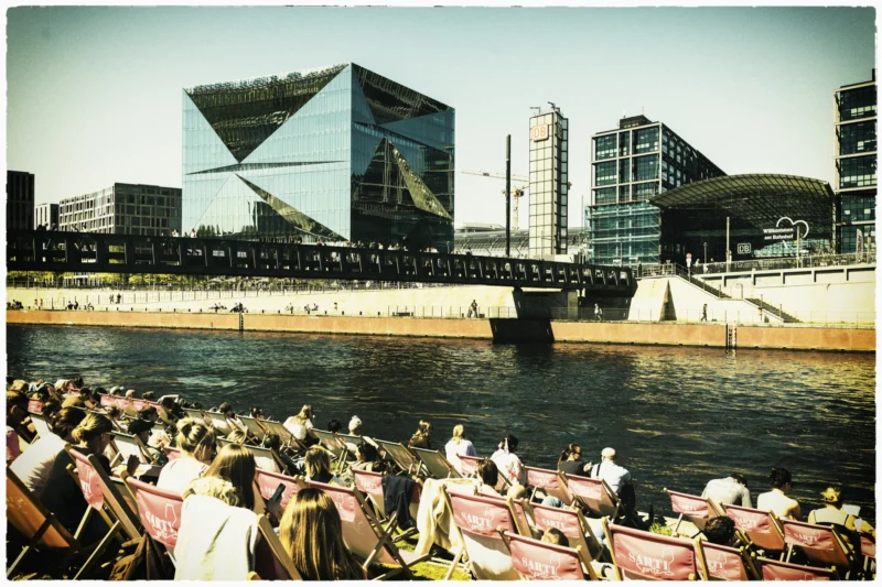 Cette photographie montre une foule assise en bordure d’un plan d’eau, regardant vers un pont et des bâtiments modernes. Le sujet principal est l’événement public, probablement un spectacle ou une projection sur les structures urbaines.La composition est dynamique, avec un cadrage large incluant le public, l’eau et les architectures futuristes. La lumière est douce, typique d’un jour ensoleillé, créant des contrastes nets. Les couleurs dominantes sont des tons chauds (beiges, bruns) et des teintes froides (bleu de l’eau, gris des bâtiments).Le style évoque un mélange de photographie documentaire et artistique, avec une ambiance conviviale et moderne.