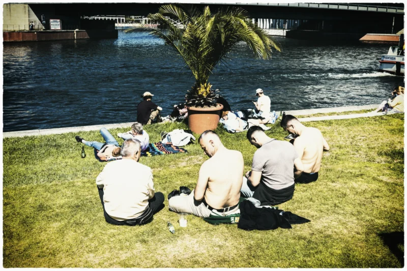 Cette photographie montre un groupe de personnes en tenue blanche, assises ou allongées sur une pelouse près d’un plan d’eau. Le sujet principal semble être ce rassemblement informel, avec une composition centrée sur eux.Le cadrage inclut un arbre en pot et un pont en arrière-plan. La lumière est naturelle, probablement en journée, avec des ombres marquées. Les couleurs dominantes sont les verts de la pelouse, les tons blancs des vêtements et les bleus du ciel et de l’eau.Le style est documentaire et spontané, évoquant une ambiance détendue et conviviale.