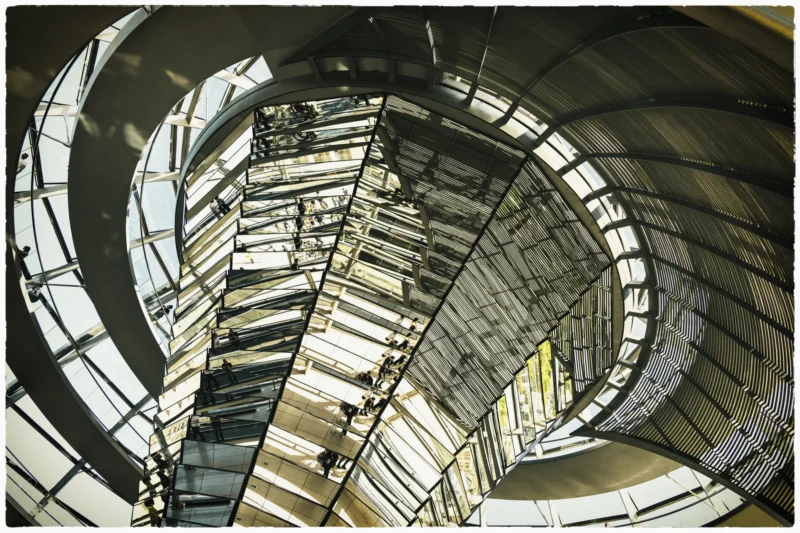 Cette photographie met en scène une structure architecturale moderne en forme d’arcs métalliques et de panneaux réfléchissants. Le sujet principal est l’assemblage complexe de ces éléments, formant des courbes dynamiques.La composition joue sur les lignes courbes et les jeux de symétrie, renforcés par un cadrage serré. La lumière, naturelle et diffuse, accentue les reflets et les contrastes entre ombres et surfaces brillantes. Les couleurs dominantes sont les tons froids (gris, argent) et quelques touches de beige.Le style est industriel et contemporain, évoquant une ambiance futuriste et presque abstraite, mêlant rigueur géométrique et mouvement.