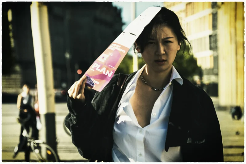 Cette photographie met en scène une jeune femme comme sujet principal, vêtue d’un costume blanc et d’une veste noire, tenant une grande glace à main. La composition est simple et centrée sur elle, avec un arrière-plan flou montrant une rue urbaine.Le cadrage est serré, mettant l’accent sur son visage et ses gestes. La lumière semble naturelle, avec des contrastes doux, créant une ambiance légèrement dramatique. Les couleurs dominantes sont les tons neutres (blanc, gris, noir) avec une touche de rose sur la glace.Le style évoque un réalisme contemporain, tandis que l’ambiance, à la fois sobre et intense, suggère une réflexion ou une introspection.