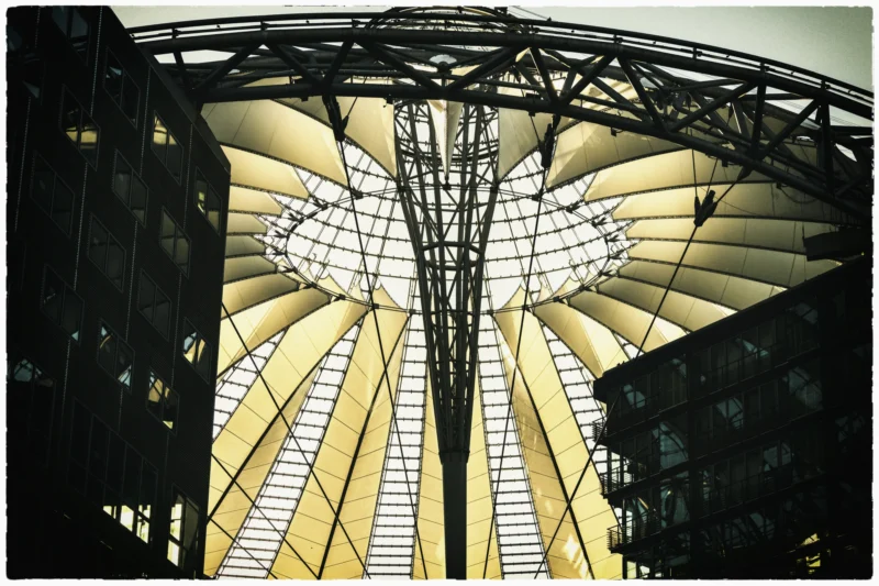 Cette photographie met en scène une structure architecturale impressionnante, probablement une gare ou un hall couvert. Le sujet principal est une immense verrière en forme de voûte, soutenue par une charpente métallique élégante.La composition est symétrique, avec des lignes convergentes vers le centre. Le cadrage est serré, mettant en valeur la structure. La lumière, naturelle et tamisée, traverse les vitraux, créant des jeux d'ombres et de clarté. Les couleurs dominantes sont des tons neutres (gris, beige) avec des éclats dorés.Le style est épuré et industriel, évoquant une ambiance à la fois moderne et historique. L'atmosphère est à la fois majestueuse et apaisante.
