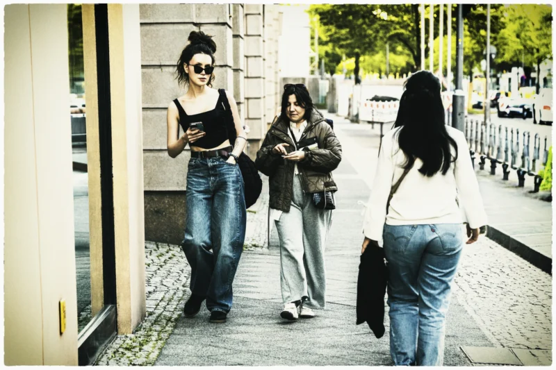 Cette photographie montre trois jeunes femmes en mouvement sur un trottoir urbain. Le sujet principal semble être leur interaction, avec une composition dynamique et naturelle.Le cadrage est serré, capturant les détails de leurs tenues et expressions. La lumière est douce, légèrement dorée, suggérant un moment en fin de journée. Les couleurs dominantes sont des tons neutres (beige, gris) avec des éclats de bleu et de blanc.Le style est documentaire et spontané, évoquant une ambiance décontractée et moderne dans un cadre urbain.