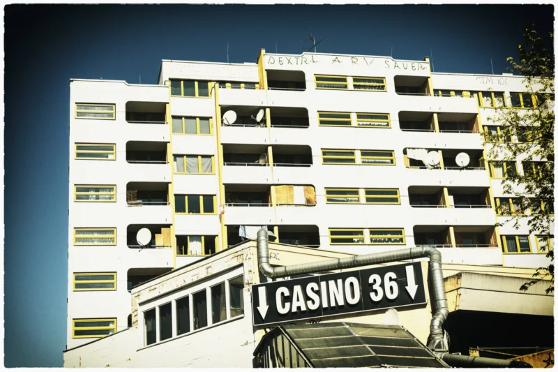 Cette photographie met en scène le **Casino 36**, bâtiment imposant aux lignes architecturales géométriques. Le **sujet principal** est l’enseigne lumineuse du casino, encadrée par une structure en arc de cercle.La **composition** est symétrique, avec un **cadrage serré** sur le bâtiment. La **lumière**, vive et contrastée, accentue les ombres et les contours, typique d’un style rétro ou vintage. Les **couleurs dominantes** sont des tons sépia et blancs, renforçant l’ambiance nostalgique.L’**ambiance** est à la fois moderne et rétro, évoquant une époque révolue avec un charme désuet.