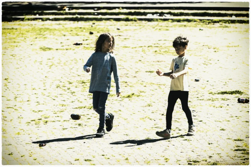 Cette photographie montre deux enfants comme sujets principaux, marchant sur une surface sableuse. La composition est simple et naturelle, avec un cadrage serré sur les enfants, centrés dans l'image.La lumière semble douce et naturelle, typique d’un jour ensoleillé. Les couleurs dominantes sont des tons neutres et naturels : beige, gris clair et quelques touches de bleu et vert.Le style est sobre et spontané, évoquant une ambiance décontractée et joyeuse, presque nostalgique grâce au filtre sépia appliqué.