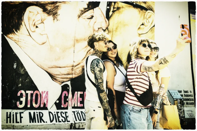 Cette photographie montre un mural représentant deux hommes s'embrassant passionnément. Le sujet principal est l'expression d'affection et de tendresse entre eux.La composition est serrée, centrée sur les visages et les corps en contact. Le cadrage est rapproché, mettant en valeur les détails des expressions et des tatouages. La lumière semble douce et naturelle, avec des contrastes atténués.Les couleurs dominantes sont des tons chauds et sépia, avec des touches de rouge et de bleu. Le style est réaliste et graphique, évoquant une ambiance à la fois intime et puissante, renforcée par le texte en cyrillique et allemand ("ЭТОН МИР" et "HILF MIR DIESE TOD").