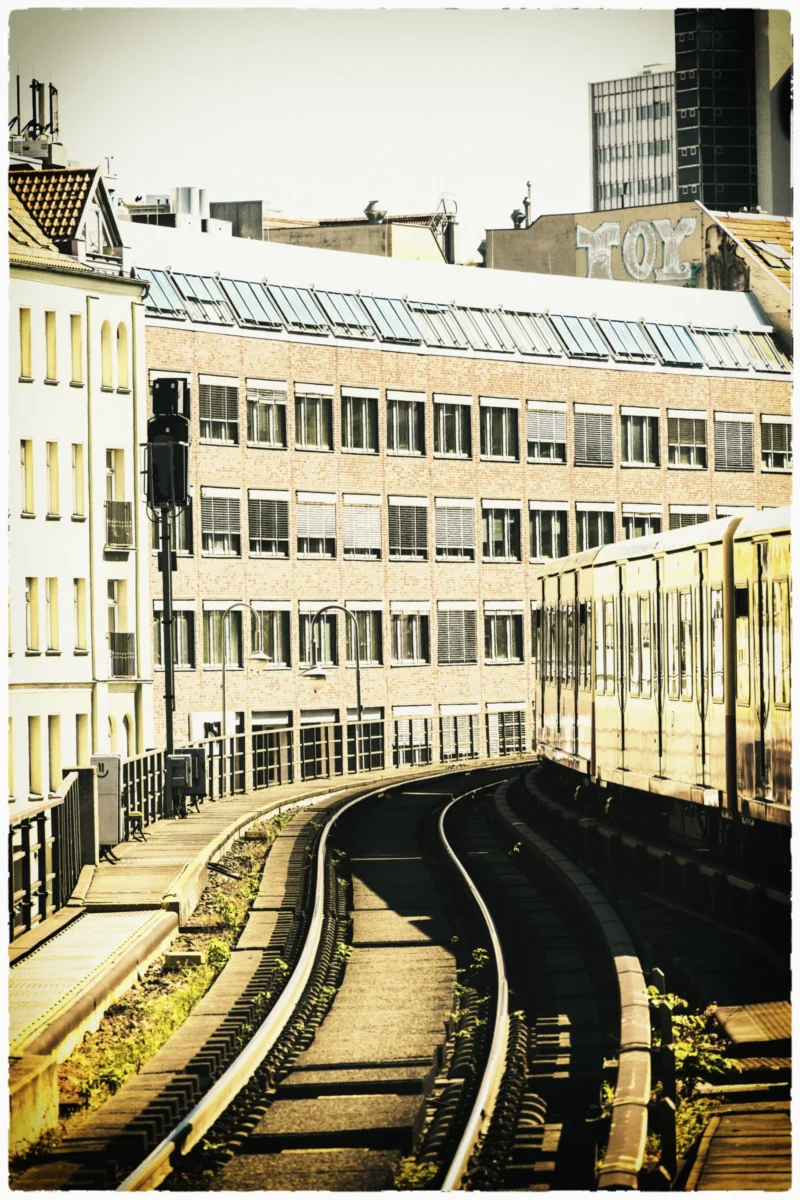 Cette photographie met en scène une voie ferrée en courbe au premier plan, encadrée par des rails et des bâtiments urbains. La composition joue sur la perspective, avec des lignes convergentes menant vers l’horizon.Le cadrage est serré, capturant une ambiance urbaine et industrielle. La lumière, douce et légèrement voilée, donne un aspect nostalgique. Les couleurs dominantes sont des tons sépia et gris, renforçant une esthétique vintage.Le style, à la fois réaliste et légèrement flou, évoque une ambiance mélancolique et intemporelle, typique des clichés anciens.
