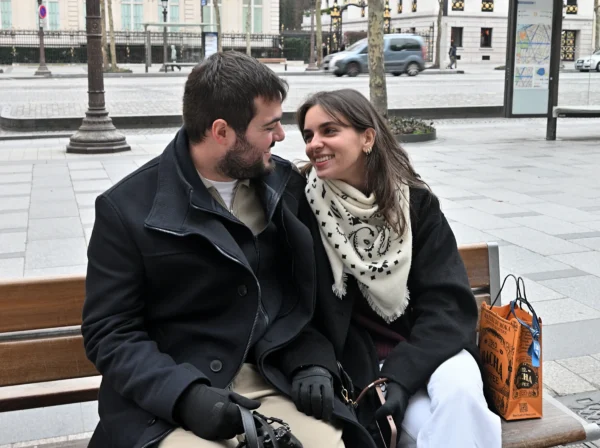 Mary · 14/02/2026 · 2784 x 2079 Cette photographie montre un couple assis sur un banc en extérieur. Le sujet principal est leur échange affectueux et complice.La composition est simple et centrée, avec un cadrage serré sur les deux personnes. L’arrière-plan flou met l’accent sur eux. La lumière semble naturelle, probablement en journée, avec des tons doux et une ambiance sobre.Les couleurs dominantes sont les teintes neutres : gris, noir et beige, adoucies par les détails chauds comme le pull blanc et le foulard. Le style est réaliste et spontané, évoquant une ambiance intime et chaleureuse dans un cadre urbain.