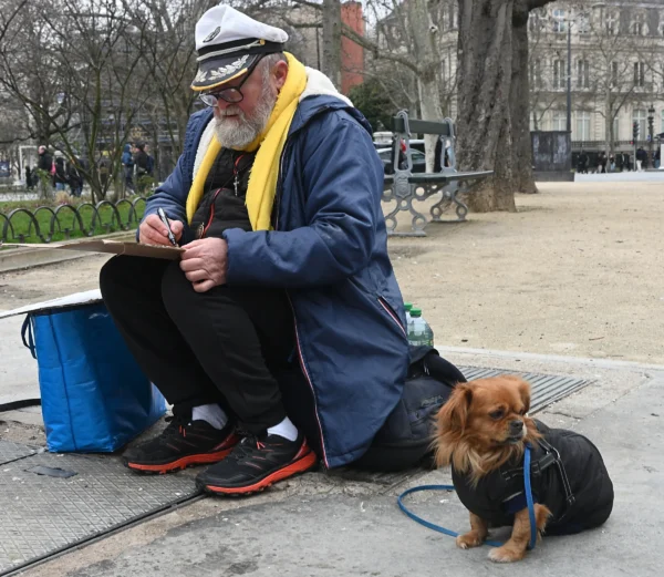 Mary · 14/02/2026 · 2784 x 2418 Cette photographie met en scène un homme assis en plein air, vêtu d’un chapeau blanc et d’un manteau bleu, jouant de la guitare. Son chien, un petit caniche, est attaché à ses pieds.La composition est naturelle, avec un cadrage serré sur les deux sujets. L’arrière-plan montre un parc urbain avec des arbres et des bâtiments.La lumière semble douce, probablement naturelle, créant des ombres légères. Les couleurs dominantes sont les tons froids (bleu, gris) et chauds (marron du chien, jaune du manteau).Le style est documentaire, capturant une scène de vie quotidienne. L’ambiance est paisible et authentique.