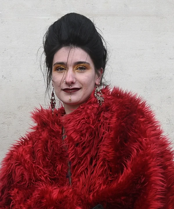 Mary · 14/02/2026 · 2331 x 2784 Cette photographie met en scène une personne comme sujet principal, vêtue d’un long manteau en fourrure rouge vif. Le cadrage est serré, centré sur son visage et sa tenue, avec un fond neutre et flou.La lumière semble naturelle et douce, éclairant le visage sans ombres marquées. Les couleurs dominantes sont le rouge éclatant du manteau et les tons neutres du fond.Le style évoque un mélange de théâtralité et de singularité, renforcé par le maquillage sombre et la coiffure volumineuse. L’ambiance est à la fois mystérieuse et audacieuse.