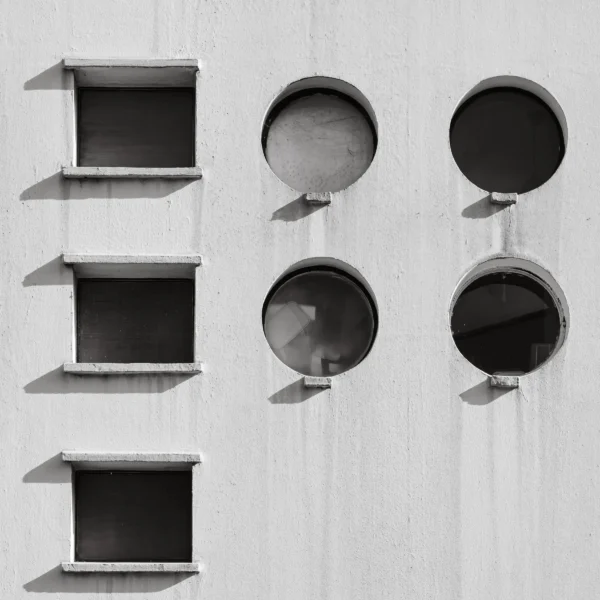 Jean-Marc · 21/02/2026 · 3041 x 3041 Cette photographie montre une façade minimaliste avec des ouvertures architecturales géométriques. Le sujet principal est une série de trois niches rectangulaires vides et de quatre cylindres circulaires, également vides, alignés verticalement.La composition est épurée et symétrique, avec un cadrage serré. La lumière naturelle crée des ombres nettes, accentuant les formes. Les couleurs dominantes sont le noir et blanc, renforçant un style moderne et abstrait. L’ambiance est sobre, presque austère, évoquant une architecture contemporaine et industrielle.