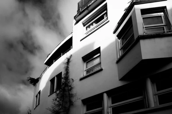 Xavier · 1600 x 1067 Cette photographie en noir et blanc met en scène un immeuble moderne aux lignes géométriques marquées. Le sujet principal est l’architecture, avec ses balcons et ses fenêtres alignées. La composition est dynamique, avec un cadrage bas mettant en valeur la verticalité du bâtiment.La lumière, contrastée, accentue les ombres et les formes, créant un effet dramatique. Les couleurs dominantes, bien que monochromes, jouent sur les nuances de gris et de noir, renforçant l’ambiance sombre et mystérieuse. Le style est épuré et contemporain, évoquant une atmosphère à la fois urbaine et mélancolique.