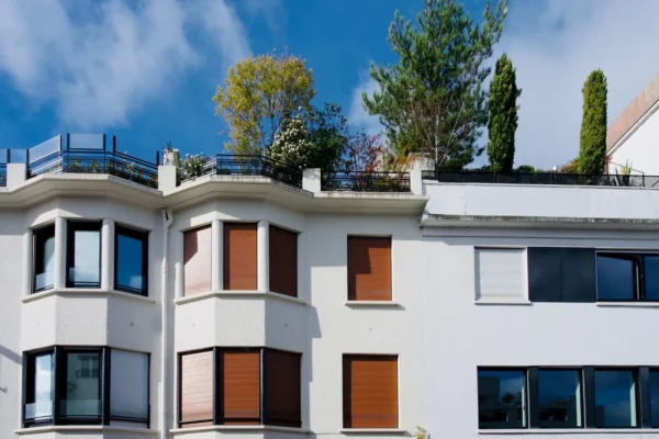 Xavier · 1600 x 1067 Cette photographie met en scène un immeuble moderne aux lignes épurées. Le sujet principal est la façade blanche aux grandes fenêtres et balcons en bois orangé, surmontée d’une terrasse végétalisée.La composition est symétrique et verticale, avec un cadrage serré sur l’architecture. La lumière, naturelle et douce, souligne les contrastes entre les surfaces claires et les teintes boisées. Les couleurs dominantes sont le blanc, l’orange et le vert des plantes.Le style est contemporain et minimaliste, évoquant une ambiance calme et urbaine, où la nature s’intègre harmonieusement à l’architecture.