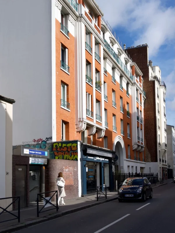 Xavier · 1201 x 1600 Cette photographie montre une rue urbaine avec un immeuble en façade principale. Le sujet principal est un bâtiment de style haussmannien aux teintes orangées et blanches, avec des balcons en fer forgé.La composition est équilibrée, avec un cadrage serré sur l’angle de la rue. La lumière est naturelle, éclairant les façades sans ombre marquée, suggérant un jour ensoleillé. Les couleurs dominantes sont les tons beiges, blancs et orangés, avec des touches de bleu et de vert sur les enseignes.L’ambiance est typiquement parisienne, calme et élégante, reflétant le charme des quartiers historiques.