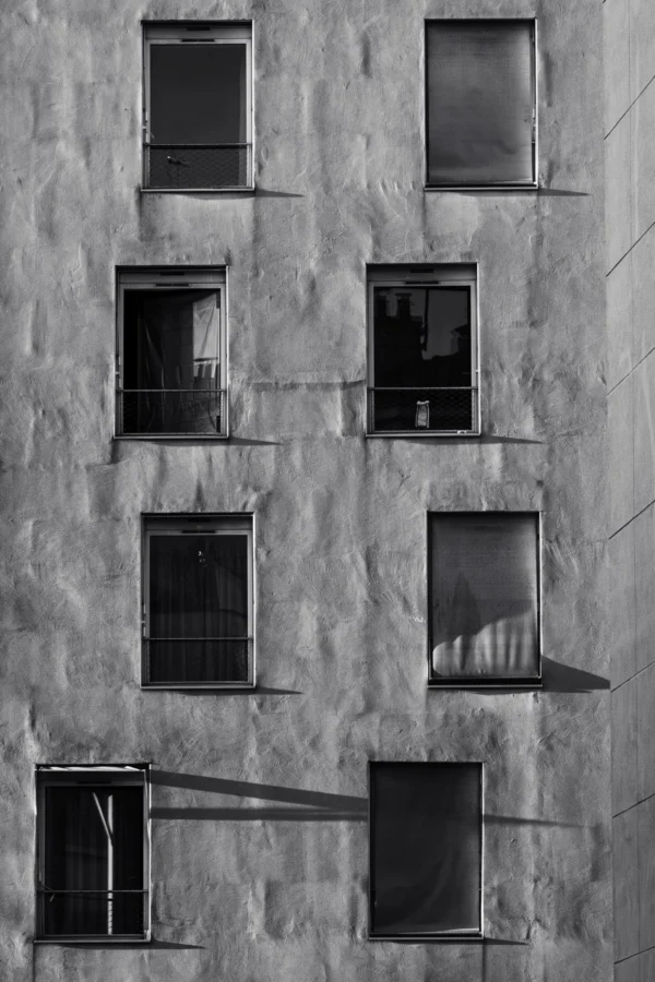 Daniel · 1621 x 2432 Cette photographie met en scène une façade d’immeuble en pierre grise, aux fenêtres rectangulaires alignées. Le sujet principal est l’architecture brute et les jeux d’ombres.La composition est symétrique et géométrique, avec un cadrage serré sur les fenêtres. La lumière, naturelle et contrastée, accentue les textures et crée des ombres marquées. Les couleurs dominantes sont les gris et noirs, renforçant une ambiance austère et minimaliste.Le style est documentaire et épuré, évoquant une esthétique industrielle. L’ambiance est froide et contemplative, presque mélancolique.
