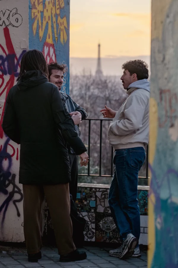 Daniel · 3155 x 4733 Cette photographie montre trois hommes discutant près d’une rambarde métallique. Le sujet principal est leur interaction, avec un cadrage serré sur eux.La composition place les personnages en premier plan, tandis qu’un mur tagué et la Tour Eiffel en arrière-plan donnent une impression d’urbanité. La lumière est douce, typique d’un coucher de soleil, créant une ambiance chaleureuse et tamisée.Les couleurs dominantes sont les tons froids (bleu, gris) contrastant avec les touches chaudes (jaune du ciel). Le style est documentaire et naturel, évoquant une scène de vie quotidienne dans un cadre parisien. L’ambiance est détendue et légèrement mélancolique.