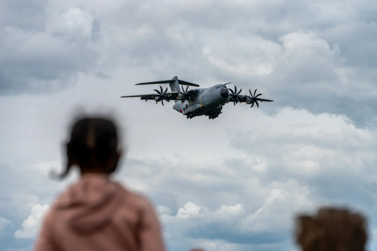 Marc · 08/06/2025 · 7008 x 4672 Cette photographie met en scène un avion militaire en vol comme sujet principal. La composition montre l’appareil en arrière-plan, légèrement flou, avec des silhouettes de personnes en premier plan, dont une enfant en rouge.Le cadrage est serré sur les observateurs, créant un contraste entre leur proximité et la distance de l’avion. La lumière est naturelle, avec des nuages dispersés, apportant une ambiance claire mais légèrement dramatique. Les couleurs dominantes sont les teintes grises et bleutées du ciel, rehaussées par les tons chauds des vêtements des personnes. Le style est documentaire, capturant un moment authentique et immersif.