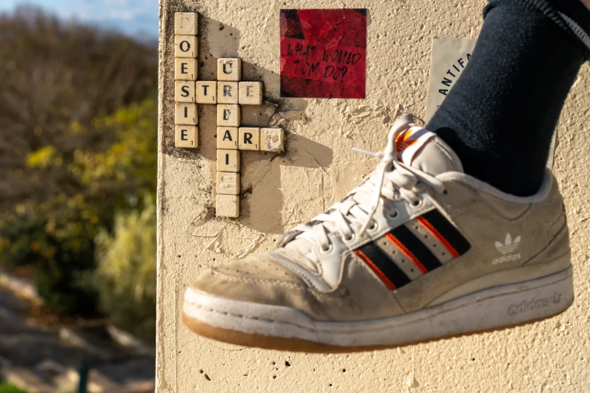 Marc · 30/11/2025 · 7008 x 4672 Cette photographie met en scène une **chaussure Adidas** (modèle Stan Smith) comme sujet principal, posée sur un mur en béton. Le cadrage divise l’image : à gauche, une vue floue de nature (végétation, chemin), et à droite, un détail du pied et de la chaussure.La lumière est naturelle, douce et légèrement contrastée, créant des ombres marquées. Les **couleurs dominantes** sont les tons neutres (beige, gris) de la chaussure et du mur, rehaussés par les détails rouges et blancs des bandes. L’ambiance est à la fois **urbaine et minimaliste**, avec une touche organique apportée par le fond flou. Le style est épuré et artistique.