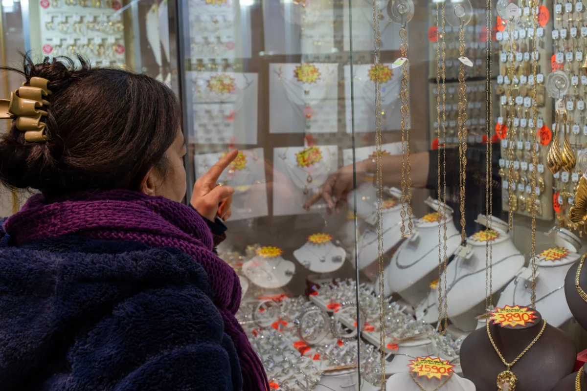 Marc · 30/11/2025 · 7008 x 4672 Cette photographie montre une femme observant des bijoux dans une vitrine. Le sujet principal est les bijoux en or exposés, notamment des boucles d’oreilles et des plateaux.La composition est serrée, mettant en valeur les détails des bijoux et l’attention de la cliente. Le cadrage est rapproché, centré sur la vitrine et la personne.La lumière est douce et naturelle, éclairant bien les objets. Les couleurs dominantes sont les tons chauds (or, brun) et quelques touches de rouge et noir.L’ambiance est artisanale et chaleureuse, évoquant un magasin de joaillerie traditionnelle.
