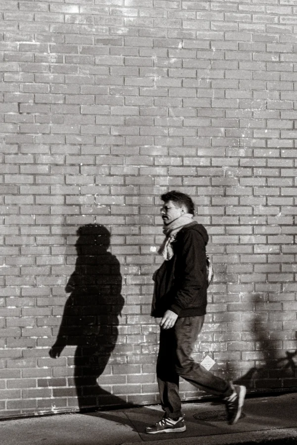 Marc · 07/02/2026 · 2494 x 3741 Cette photographie en noir et blanc met en scène deux silhouettes en mouvement devant un mur de briques. Le sujet principal semble être l’homme en premier plan, vêtu d’un manteau et d’un chapeau, marchant avec une canne. La composition joue sur les contrastes et les ombres, créant une ambiance dynamique.Le cadrage serré et la lumière latérale accentuent les contours et les jeux d’ombres. Les tons dominants sont les gris et noirs, renforçant une atmosphère à la fois urbaine et mélancolique. Le style rappelle le photojournalisme documentaire des époques classiques, évoquant une touche de nostalgie et de réalisme brut.