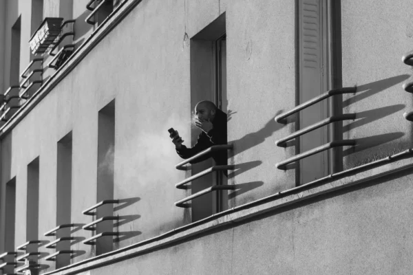 Marc · 07/02/2026 · 3251 x 2167 Cette photographie en noir et blanc met en scène une personne comme sujet principal, assise sur un balcon, absorbée par son téléphone. La composition est épurée, avec un cadrage serré sur le balcon et une partie du bâtiment.Les jeux d’ombres et de lumière, créés par les barreaux et les moulures, ajoutent du relief. Les tons dominants sont les gris et noirs, renforçant une ambiance sobre et introspective. Le style est documentaire et minimaliste, évoquant une solitude urbaine et une modernité mélancolique.