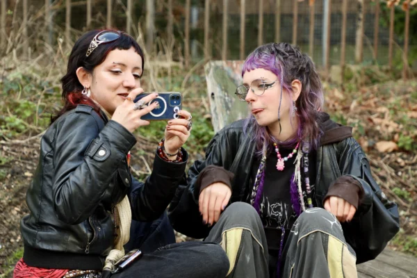 Francoise · 07/02/2026 · 6000 x 4000 Cette photographie montre deux jeunes femmes assises dans un cadre naturel boisé. Le sujet principal est leur interaction : l’une prend une photo de l’autre avec un appareil compact.La composition est symétrique et intime, avec un cadrage serré. La lumière est douce et naturelle, probablement en journée. Les couleurs dominantes sont les tons verts de la végétation, les teintes sombres des vêtements (noir, marron) et les touches vives des accessoires (violet, bleu).Le style est décontracté et spontané, évoquant une ambiance amicale et décontractée, presque bohème.