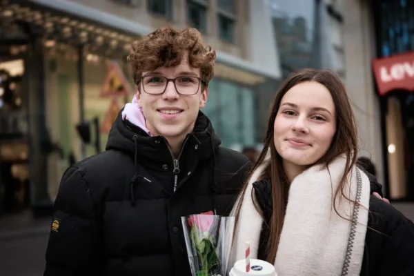 Claude · 1080 x 720 Cette photographie met en scène deux jeunes posant côte à côte. Le sujet principal est leur complicité souriante, avec un cadrage serré sur leurs visages.La composition est symétrique et équilibrée, centrée sur eux. L'arrière-plan flou révèle une rue urbaine avec des enseignes et des bâtiments.La lumière est naturelle, douce, probablement en journée. Les couleurs dominantes sont les tons froids (noirs, gris) contrastés avec les teintes chaudes de leurs vêtements (blanc, marron) et les touches de rose des fleurs.Le style est spontané et naturel, évoquant une ambiance chaleureuse et amicale.