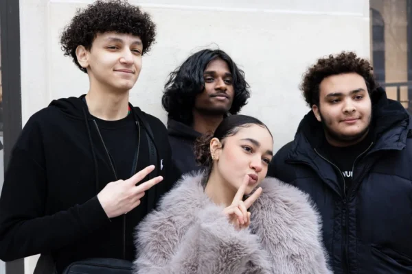 Claude · 1080 x 720 Cette photographie met en scène un groupe de quatre jeunes posant ensemble. Le sujet principal semble être la jeune femme au centre, entourée de ses amis.La composition est symétrique et serrée, avec un cadrage rapproché. La lumière est naturelle et douce, éclairant uniformément les visages. Les couleurs dominantes sont les tons neutres (noirs, gris) et quelques touches de couleurs chaudes sur les vêtements.Le style est décontracté et urbain, évoquant une ambiance amicale et moderne. L'atmosphère est joyeuse et spontanée.