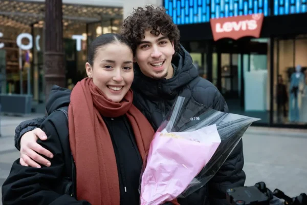 Claude · 1080 x 720 Cette photographie montre deux jeunes souriants, enlacés, posant devant un magasin. Le sujet principal est leur complicité et leur joie.La composition est simple et naturelle, avec un cadrage serré sur eux. La lumière semble douce et naturelle, probablement en journée. Les couleurs dominantes sont les tons froids (noir de leurs vêtements) et chauds (rouge du manteau de la jeune fille, teinte rose du sac).Le style est spontané et authentique, évoquant une ambiance chaleureuse et amicale en milieu urbain.
