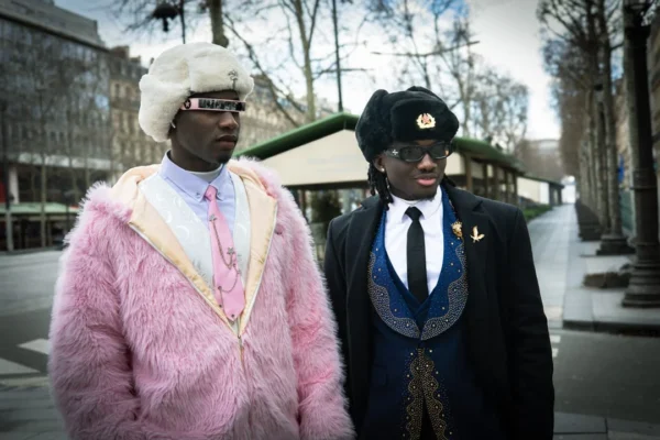 Claude · 1080 x 720 Cette photographie met en scène deux hommes comme sujets principaux, vêtus de manière excentrique : l’un en fourrure rose et chapeau blanc, l’autre en costume noir avec chapeau et décorations. La composition est symétrique et serrée, les deux personnages se faisant face.Le cadrage est serré sur eux, avec un arrière-plan flou de ville. La lumière semble naturelle, légèrement contrastée, créant des ombres douces. Les couleurs dominantes sont le rose vif, le noir et le blanc, renforçant un style théâtral et décalé. L’ambiance est à la fois mystérieuse et humoristique, évoquant une touche de fantaisie urbaine.