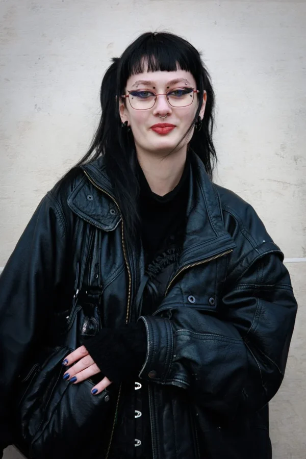 Claude · 900 x 1350 Cette photographie met en scène une personne comme sujet principal, vêtue d'une veste en cuir noire et d'un haut sombre, les bras croisés. Le cadrage est serré, centré sur elle, avec un fond neutre et lisse.La lumière semble naturelle, douce mais contrastée, mettant en valeur les traits du visage. Les couleurs dominantes sont les noirs et gris, renforçant un style sombre et élégant. L’ambiance est à la fois mystérieuse et introspective, évoquant un côté gothique ou alternatif. Le style rappelle une esthétique urbaine et moderne.