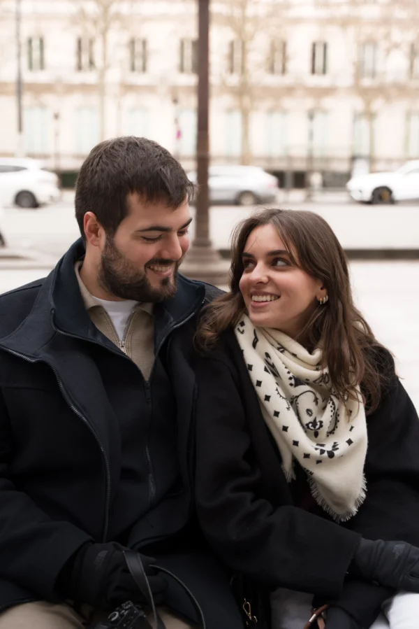 Juan · 14/02/2026 · 4774 x 7157 Cette photographie met en scène un couple assis côte à côte, souriant tendrement. Le sujet principal est leur complicité et leur proximité.La composition est simple et naturelle, avec un cadrage serré sur les visages et les épaules. L'arrière-plan flou montre une ville hivernale, avec des bâtiments et des arbres.La lumière est douce et naturelle, probablement en journée, créant des ombres légères. Les couleurs dominantes sont les tons froids (gris, noir) adoucis par les touches chaudes du manteau noir et du châle blanc de la femme.Le style est réaliste et spontané, évoquant une ambiance chaleureuse et intime.