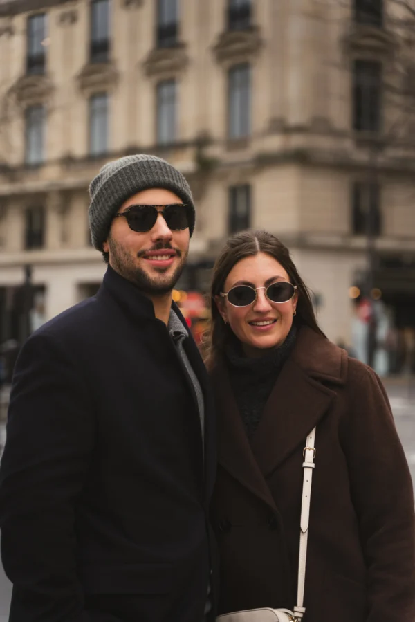 Juan · 14/02/2026 · 5304 x 7952 Cette photographie met en scène un couple souriant comme sujet principal. Le cadrage est serré, centré sur eux, avec un arrière-plan flou montrant un bâtiment classique et une rue animée.La lumière est naturelle, douce et légèrement tamisée, créant des ombres douces. Les couleurs dominantes sont les tons neutres (beige, gris) avec des touches de brun et noir dans leurs vêtements. Le style est décontracté et urbain, évoquant une ambiance moderne et chaleureuse, presque intimiste malgré le cadre extérieur.