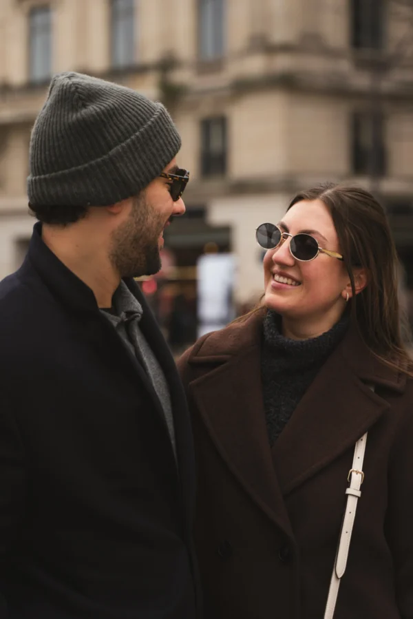 Juan · 14/02/2026 · 4315 x 6469 Cette photographie met en scène deux personnes en conversation. Le sujet principal est un homme en pull noir et bonnet, et une femme avec des lunettes teintées et une veste marron. La composition est serrée, centrée sur leurs visages et épaules, créant une ambiance intime. Le cadrage est rapproché, mettant l'accent sur leur interaction.La lumière semble naturelle, douce et légèrement tamisée, typique d'une journée urbaine. Les couleurs dominantes sont les tons neutres (noir, marron, gris) avec des détails contrastés comme les lunettes de la femme. Le style est documentaire et spontané, évoquant une ambiance urbaine et chaleureuse.