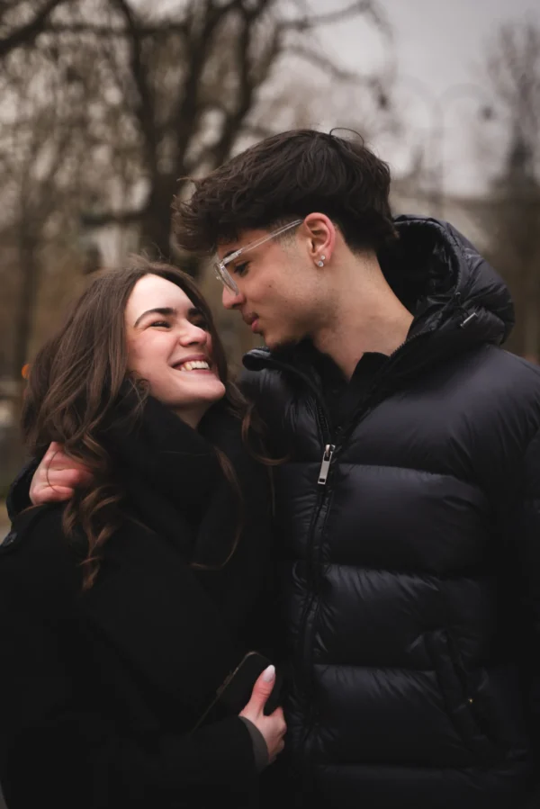 Juan · 14/02/2026 · 4674 x 7008 Cette photographie met en scène un couple en étreinte tendre. Le sujet principal est deux jeunes gens souriants, l’un avec des lunettes et une veste noire, l’autre avec des cheveux longs.La composition est serrée et centrée sur eux, avec un cadrage rapproché. L’arrière-plan flou, composé d’arbres et de bâtiments, crée un effet bokeh.La lumière est douce et naturelle, probablement en journée. Les couleurs dominantes sont les tons neutres (gris, noir) et quelques touches de vert et brun.Le style est épuré et romantique, évoquant une ambiance chaleureuse et intime.