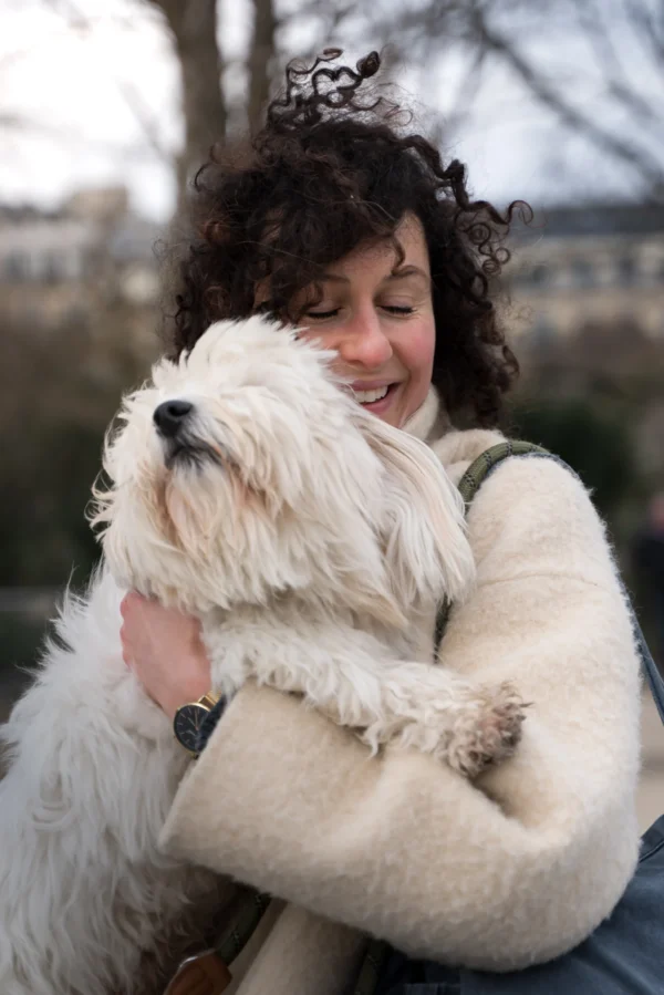 Juan · 14/02/2026 · 4057 x 6082 Cette photographie met en scène une femme comme sujet principal, souriante et câlinant un petit chien blanc à poils longs. La composition est centrée sur eux deux, avec un cadrage serré qui isole les sujets du fond flou.La lumière naturelle, douce et légèrement tamisée, adoucit les traits et crée une ambiance chaleureuse. Les couleurs dominantes sont les tons neutres (blanc du chien, beige du manteau) avec des touches naturelles et douces.Le style est simple et spontané, évoquant une scène intime et affectueuse. L’ambiance est réconfortante et pleine de tendresse.