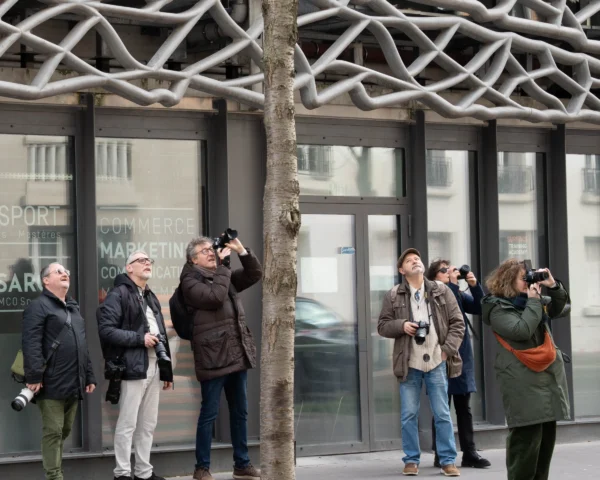 Alice · 21/02/2026 · 3187 x 2550 Cette photographie montre un groupe de cinq personnes prenant des photos devant un bâtiment moderne. Le sujet principal est l’activité photographique collective, avec des appareils variés.La composition est dynamique, centrée sur les individus en action. Le cadrage est serré, mettant en valeur les visages et les gestes. La lumière est naturelle, probablement en journée, avec des ombres douces. Les couleurs dominantes sont neutres (gris, beige) avec des touches de vert et de noir.Le style est documentaire et spontané, capturant un moment authentique. L’ambiance est studieuse et collaborative, évoquant une visite touristique ou culturelle.