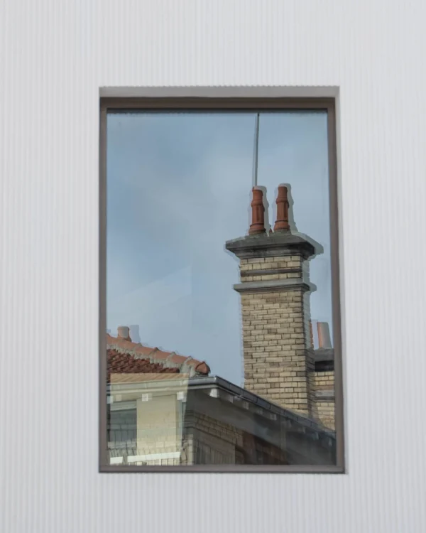 Alice · 1882 x 2353 Cette photographie montre une vue reflétée dans un cadre de fenêtre. Le **sujet principal** est une cheminée en brique avec deux pots rouges, surmontant un toit en ardoise.La **composition** est simple et épurée, centrée sur l'élément architectural. Le **cadrage** est serré, mettant en valeur uniquement la partie supérieure du bâtiment. La **lumière** est naturelle, douce et légèrement voilée, créant des ombres douces.Les **couleurs dominantes** sont les tons gris-beige des briques, les rouges des pots et les teintes bleutées du ciel reflété. Le style est **minimaliste et réaliste**, évoquant une **ambiance calme et intemporelle**.