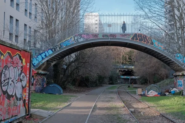 Dominique · 900 x 600 Cette photographie montre un pont couvert de graffitis, sujet principal de l'image. La composition met en valeur une voie ferrée abandonnée sous le pont, encadrée par des murs tagués et des bâtiments en arrière-plan.Le cadrage est serré, guidant le regard vers le centre où un banc et des déchets sont visibles. La lumière naturelle, douce et légèrement bleutée, crée une ambiance sobre et mélancolique. Les couleurs dominantes sont les tons froids (bleus, gris) contrastant avec les éclats de rouge et de bleu des graffitis. Le style est réaliste et documentaire, évoquant une atmosphère urbaine désolée et artistique.
