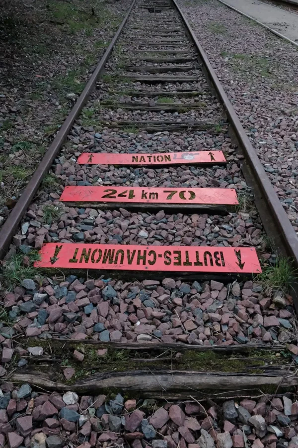 Dominique · 900 x 1350 Cette photographie montre des rails de chemin de fer avec trois panneaux rouges placés en travers. Le sujet principal est ces panneaux indiquant des distances ("Buttes-Chaumont", "Nation", "241 m" et "70").La composition est symétrique et horizontale, centrée sur les panneaux. Le cadrage est serré, mettant en avant les détails des rails et des panneaux. La lumière semble naturelle, légèrement tamisée, créant des ombres douces. Les couleurs dominantes sont le rouge vif des panneaux, le gris des rails et le brun des graviers. L’ambiance est industrielle et minimaliste, évoquant un lieu utilitaire et abandonné.