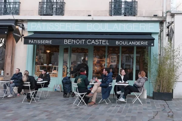 Dominique · 900 x 600 Cette photographie montre l'extérieur d'une pâtisserie-boulangerie nommée **Benoît Castel**. Le sujet principal est un groupe de personnes assises à des tables en terrasse, discutant et profitant de leur café ou pâtisserie.La composition est équilibrée, avec une perspective légèrement en contre-plongée. Le cadrage inclut une partie de la rue et les façades des bâtiments. La lumière est naturelle, douce, et semble provenir d’un ciel légèrement nuageux. Les couleurs dominantes sont les tons pastel (bleu clair, vert) et les neutres (beige, gris).L’ambiance est détendue et conviviale, typique d’un café parisien. Le style est réaliste et quotidien.