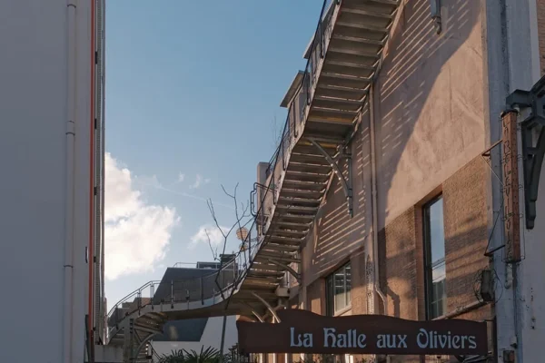 Dominique · 1000 x 667 Cette photographie met en scène un bâtiment avec une enseigne "La Faille aux Ouvriers". Le sujet principal est l’architecture industrielle, marquée par des murs en brique et une structure métallique en escalier extérieur.La composition joue sur les lignes verticales et horizontales, créant une perspective dynamique. Le cadrage inclut partiellement un ciel dégagé, ajoutant de la profondeur.La lumière naturelle, douce et légèrement dorée, évoque un moment en fin de journée. Les couleurs dominantes sont les tons beiges des briques, contrastant avec le bleu du ciel. L’ambiance est à la fois urbaine et chaleureuse, mêlant modernité et nostalgie.
