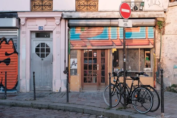 Dominique · 1000 x 666 Cette photographie met en scène une boutique aux allures vintage, avec une façade en bois et des enseignes colorées. Le sujet principal est l’entrée de ce magasin, encadrée par des portes vitrées ouvertes.La composition est symétrique, avec un vélo noir attaché devant, ajoutant une touche urbaine. Le cadrage est serré, centré sur la boutique et son environnement immédiat. La lumière est naturelle, douce et légèrement tamisée, créant des ombres douces.Les couleurs dominantes sont les tons chauds (bois, orange) et froids (bleu, gris). Le style est street photography, réaliste et authentique. L’ambiance est à la fois chaleureuse et un peu mélancolique, évoquant un quartier vivant et artistique.
